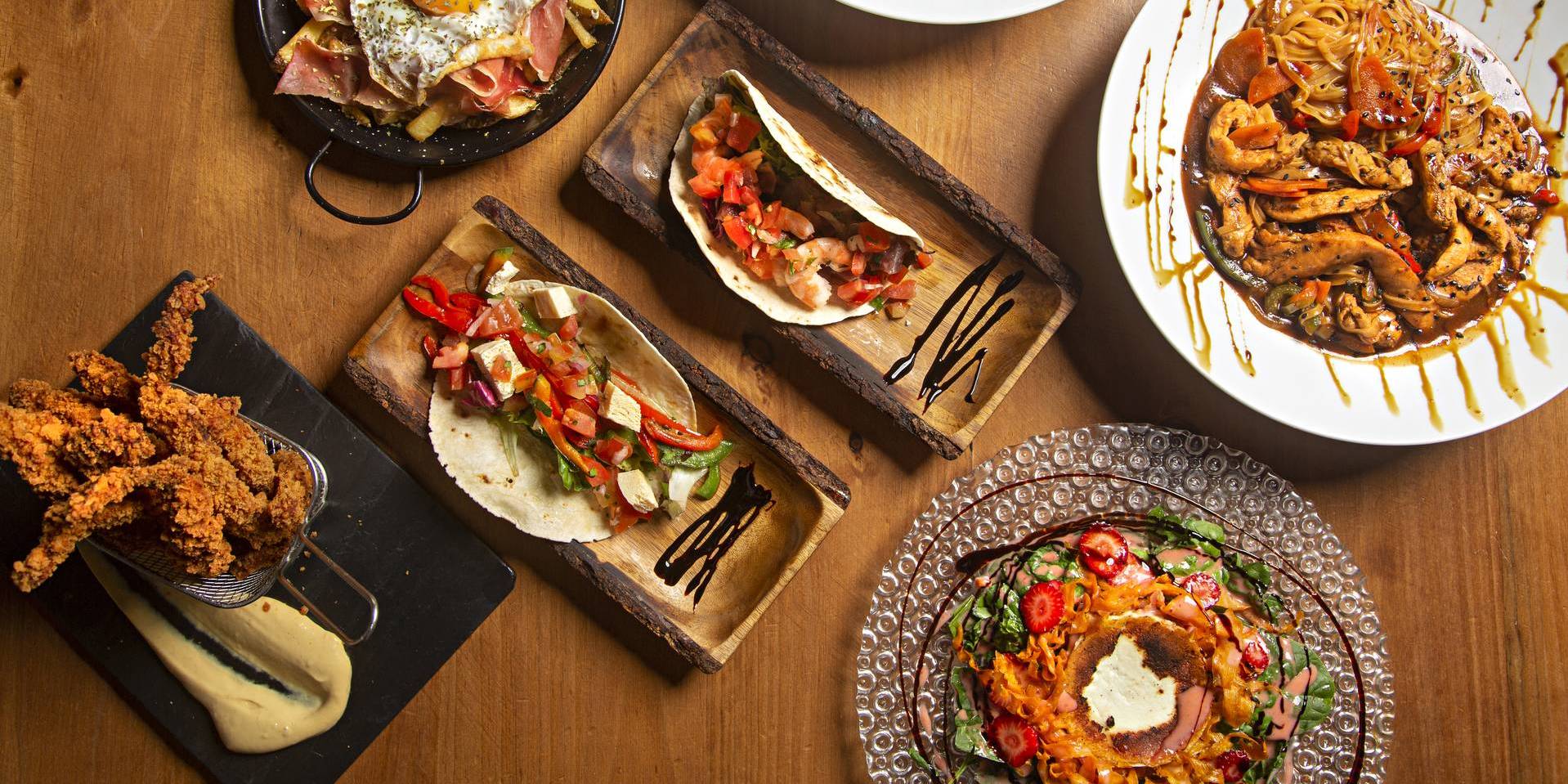 Varied food dishes of the world on wooden table, eggs, salad, chicken, tacos.